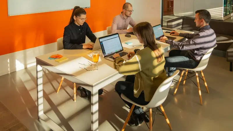 Quatre collègues travaillant sur des ordinateurs portables autour d’une table blanche dans un bureau moderne au mur orange