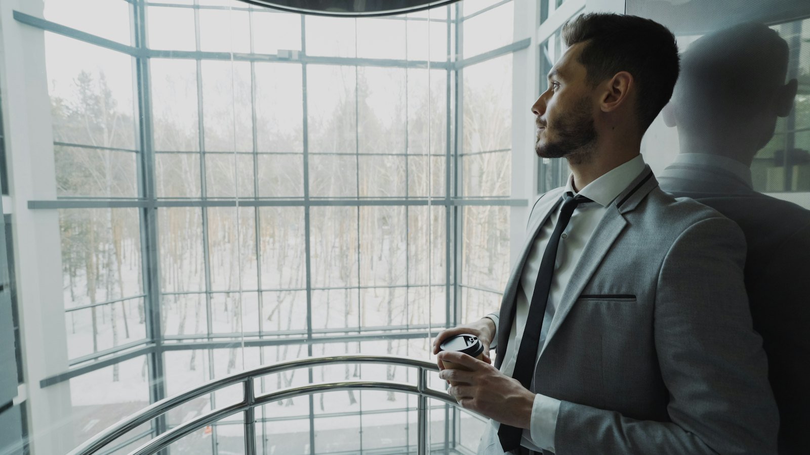 Executive in a suit holding a coffee, looking out alone through a large window in a modern office building